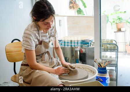 young ceramist on the lathe or potter's wheel working and shaping clay inside a pottery studio, creating creative shapes with tools. natural light Stock Photo