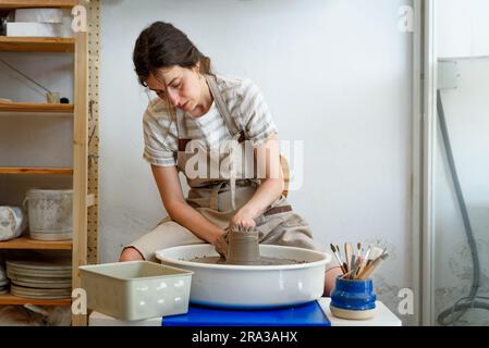 young ceramist on the lathe or potter's wheel working and shaping clay inside a pottery studio, creating creative shapes with tools. natural light Stock Photo