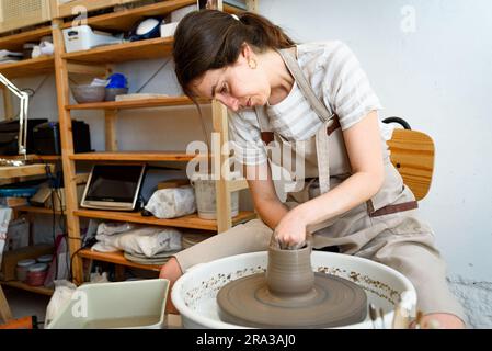 young ceramist on the lathe or potter's wheel working and shaping clay inside a pottery studio, creating creative shapes with tools. natural light Stock Photo