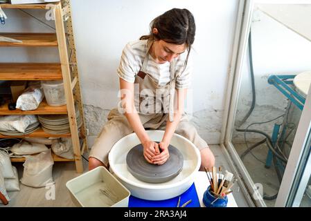 young ceramist on the lathe or potter's wheel working and shaping clay inside a pottery studio, creating creative shapes with tools. natural light Stock Photo