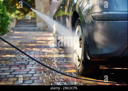 Close-up of car washing process. Unknown man directs a hose with jet of water at the rear wheel of his car, washes the vehicle in the backyard of the Stock Photo