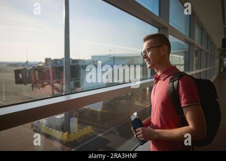 Smiling man travel by airplane. Passenger looking through window at airport runway while holding wallet with boarding pass in his hand. Stock Photo