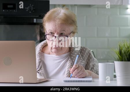 Elderly woman in glasses working using laptop at home, looking on computer Stock Photo