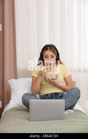 Teenage girl with headphones eating popcorn while using laptop on bed at home Stock Photo