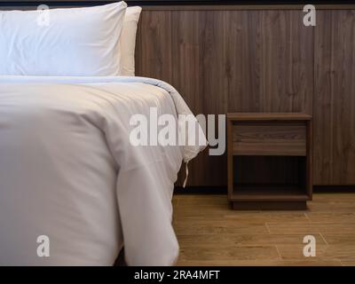 Empty wooden bedside cupboard near the bed with white blanket decorated in a hotel room. Walnut wood bed side table on teak wood tiles and brown wall Stock Photo