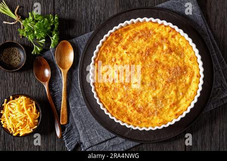 Cheddar Cheese Grits Casserole in baking dish on dark wooden table with wood spoons, fresh parsley and shredded cheese, horizontal view from above, fl Stock Photo