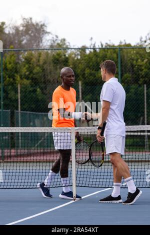 Diverse male tennis players shaking hands on outdoor court Stock Photo