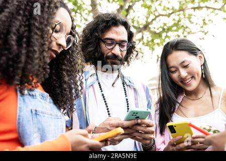 Multiethnic group of friends texting on their smartphones in the street.Three diverse young people smiling and using their phones outside. Stock Photo