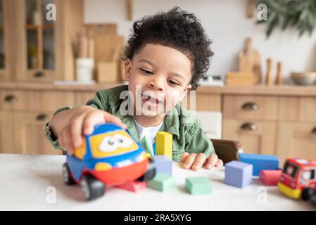 Happy little african american boy with bushy curly hair playing with colorful wooden blocks, enjoying table games playing alone at home kitchen Stock Photo