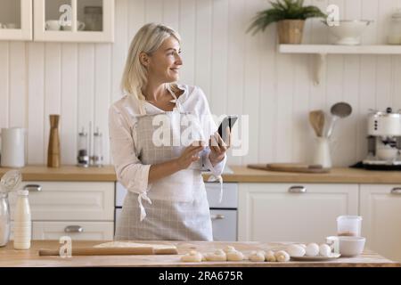 Happy pretty blonde mature housewife in apron browsing cooking recipe Stock Photo