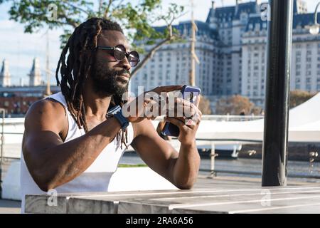 young man of African ethnicity, with dreadlocks and sunglasses, sitting outdoors touching the mobile screen browsing the internet, viewing notificatio Stock Photo