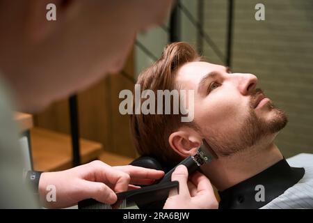 Master cuts the beard of a young client in abarbershop Stock Photo