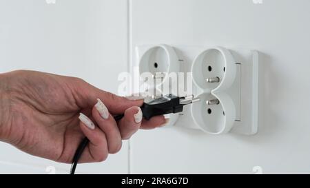 Close-up of a female hand inserting a black electric plug into a double European electrical socket isolated on a white wall. Renovation in a new apart Stock Photo