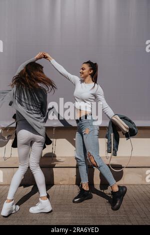 Two pretty brunette girls dancing together in front of the grey wall Stock Photo