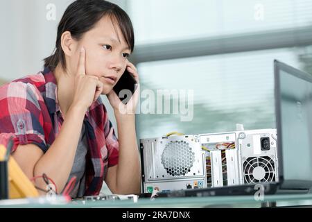 female technician talking on smartphone and looking at laptop screen Stock Photo