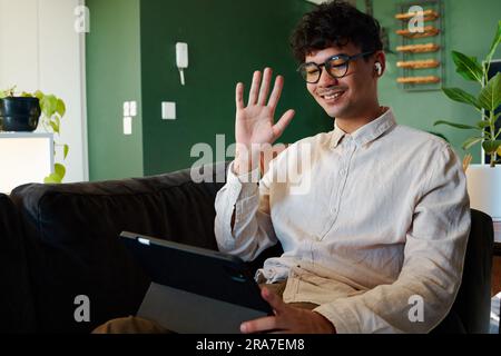 Young multiracial man smiling while using digital tablet and waving on sofa at home Stock Photo