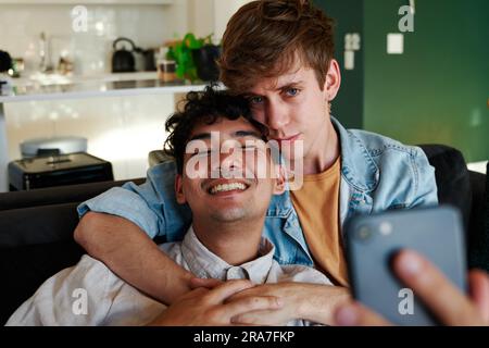 Young gay couple taking selfie with mobile phone while embracing on sofa at home Stock Photo