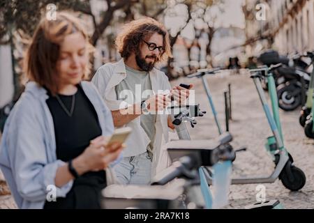 Couple rent electric push scooters using mobile phones while standing on street in city Stock Photo