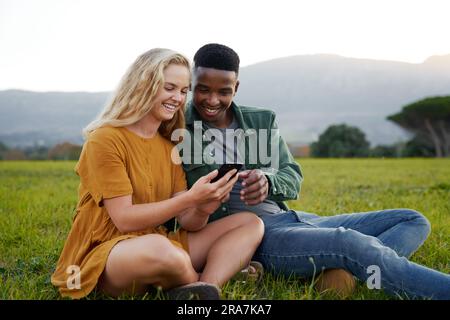 Happy multiracial young couple sitting and using mobile phone in field Stock Photo