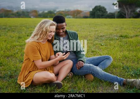 Happy multiracial young couple sitting and smiling while using mobile phone in field Stock Photo