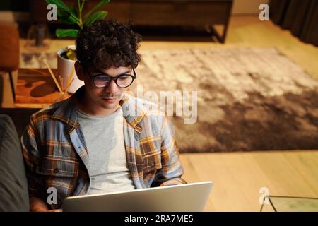 Focused young multiracial man wearing eyeglasses using laptop on sofa at home Stock Photo