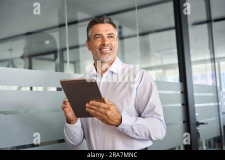 Happy mid aged business man manager looking away in office using digital tablet. Stock Photo