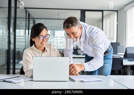 Mature boss manager helping Asian employee working on laptop in office. Stock Photo