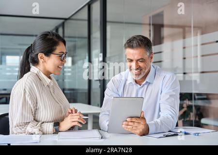 Two happy diverse executives discussing work using digital tablet in office. Stock Photo
