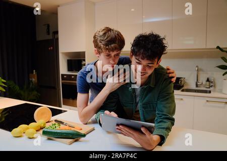 Young gay couple in casual clothing smiling while using digital tablet next to food in kitchen Stock Photo