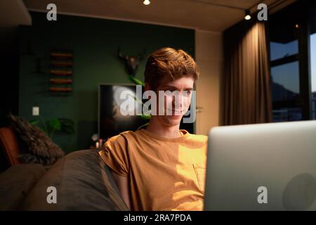 Happy young caucasian man wearing casual clothing smiling while using laptop on sofa at home Stock Photo