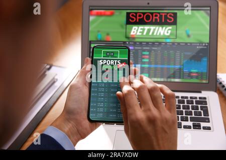 Man betting on sports using smartphone and laptop at table, closeup. Bookmaker websites on displays Stock Photo