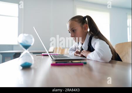 Caucasian girl sits at a desk at school and studies at a laptop. Hourglass on the table. Stock Photo