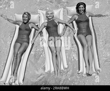 United States:  c. 1962 Three young women in matching one piece bathing suits floating together on rafts in a pool. Stock Photo