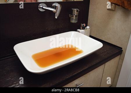 The rusty water gushes out of the faucet, making the sink wet and dirty. The unclean water flowing from the rusty faucet in the bathroom is unfit for Stock Photo