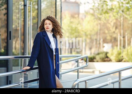 Trendy young woman in wool coat and trousers posing near modern building Stock Photo