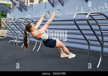 Woman doing pull-ups on rings spreading her arms to sides outdoors Stock Photo