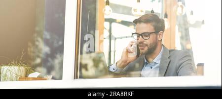 Young handsome businessman wearing eyeglasses talking by phone while working remotely in cafe Stock Photo