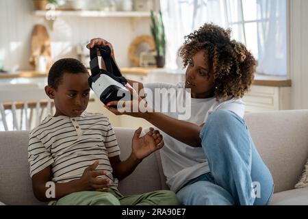 African American mom sitting with child son on sofa taking off his vr glasses, limiting screen time Stock Photo