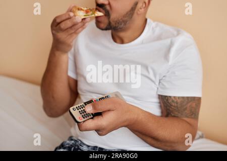 Closeup of tattooed man eating pizza at home and holding remote control while enjoying relaxing weekend Stock Photo