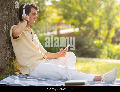 Positive millennial man chilling under tree at park, using phone Stock Photo