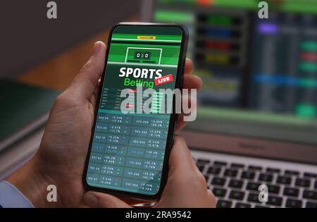 Man betting on sports using smartphone and laptop at table, closeup. Bookmaker websites on displays Stock Photo
