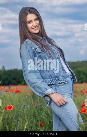 Smiling long haired  brunette girl dressed in blue jeans is posing in red poppies meadow. Vertically. Stock Photo