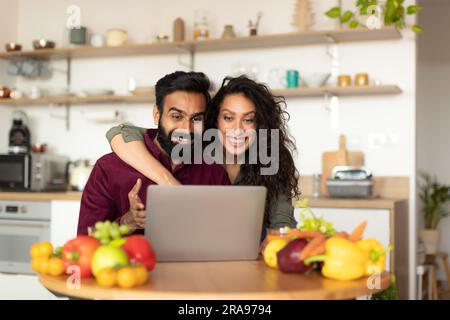 Happy arab spouses using laptop at kitchen, cheerful husband and wife shopping online or ordering food delivery Stock Photo