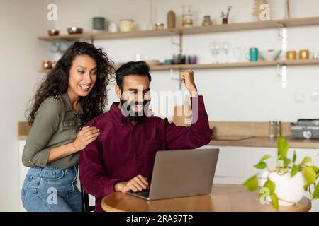 Emotional overjoyed arab spouses clenching fists, looking at laptop screen and screaming in kitchen, copy space Stock Photo