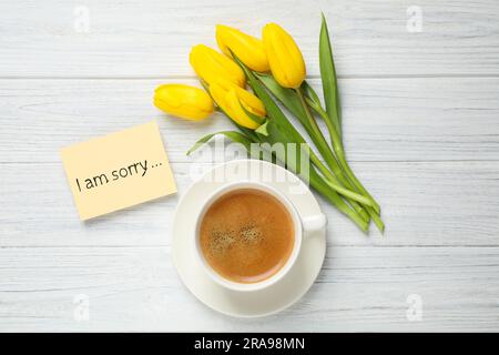 Apology. Yellow card with phrase I Am Sorry, cup of coffee and beautiful yellow tulips on white wooden table, flat lay Stock Photo