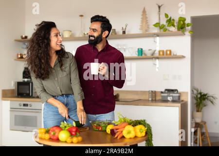 Happy young arab woman preparing healthy meal for her husband, happy man drinking tea next to his spouse cooking dinner Stock Photo