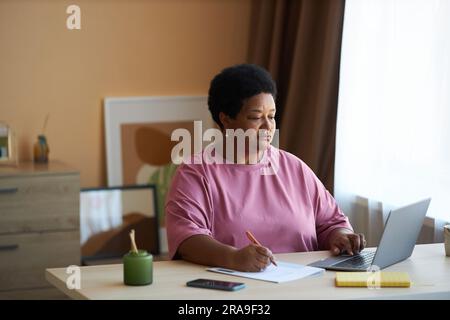 Serious senior businesswoman making notes in document while sitting by workplace in front of laptop and looking through online information Stock Photo