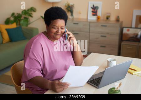 Smiling aged businesswoman with financial document talking to her employer on mobile phone while sitting by desk in front of laptop Stock Photo