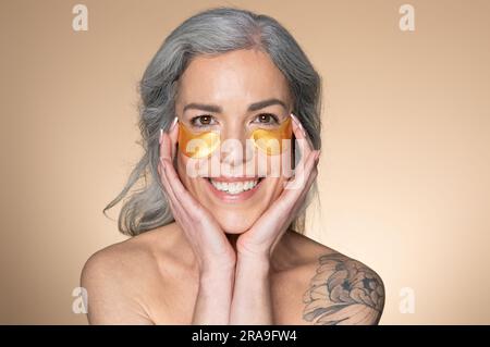 Portrait of cheerful senior woman wearing cosmetic eye patches, making rejuvenating procedure on beige studio background Stock Photo