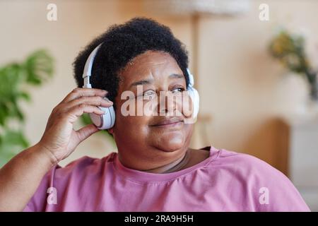 Aged restful African American woman listening to relaxing music in headphones and looking aside while sitting in front of camera Stock Photo